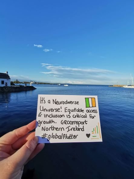 Dwylo yn dal cerdyn yn dweud “Mae'n Fydysawd niwrodiverse! Mae mynediad a chynhwysiant teg yn hanfodol ar gyfer twf. Groomsport, Gogledd Iwerddon, Hashtag GlobalFlutter. Yn y cefndir mae môr o ddŵr glas a chymylau yn yr awyr gyda thŷ gwyn i'r chwith o'r ddelwedd